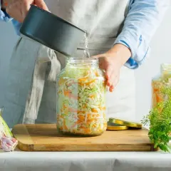 Doprawianie kiszonej kapusty. Woda z garnka zalewa słoik z kapustą i marchewką, przygotowując pyszny sok z kiszonej kapusty.